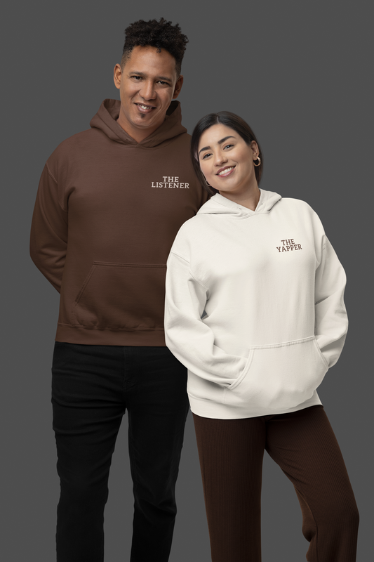 A couple posing in studio wearing matching hoodies with embroidered text – one says “THE YAPPER” and the other “THE LISTENER.”