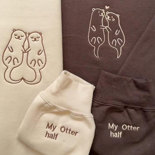 Matching couple hoodies with embroidered otter illustrations holding hands and sleeve text that reads “My Otter Half.”