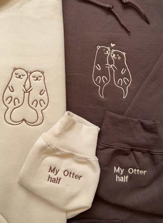 Matching couple hoodies with embroidered otter illustrations holding hands and sleeve text that reads “My Otter Half.”